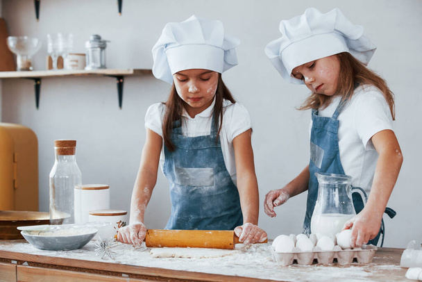 Two little girls in blue chef uniform kneading dough on the kitchen. - Foto, Imagen