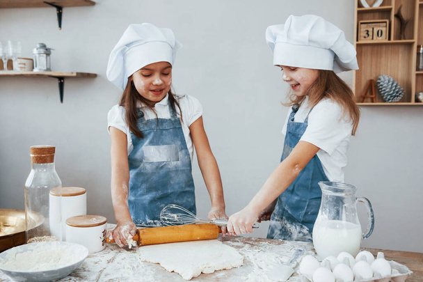 Two little girls in blue chef uniform kneading dough on the kitchen. - Foto, imagen