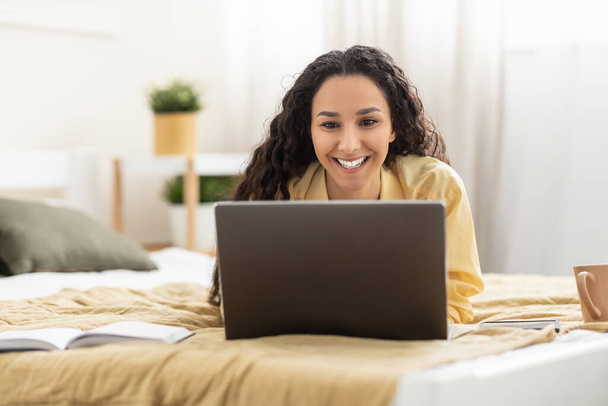 Smiling woman using laptop lying on bed at home - Fotó, kép