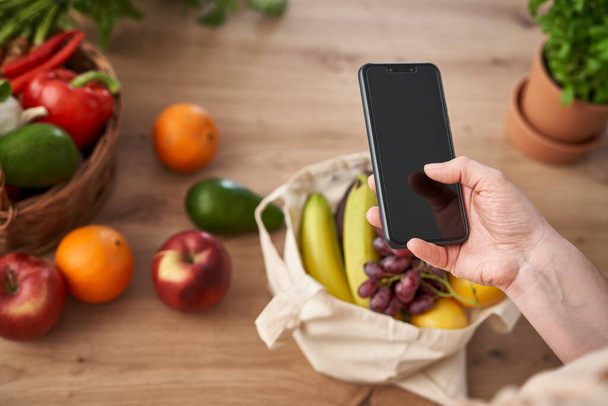 Frau benutzt Handy mit Einkaufstasche voller Obst und Gemüse - Foto, Bild