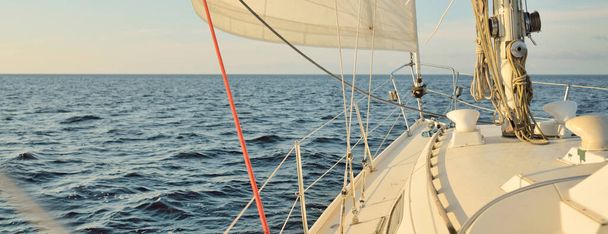 Bílá šalupa lanoví jachta plachtění na otevřeném moři při západu slunce. Jasná obloha. Pohled z paluby na příď, stěžeň, plachty. Doprava, cestování, plavba, sport, rekreace, volný čas, závodní, regata - Fotografie, Obrázek