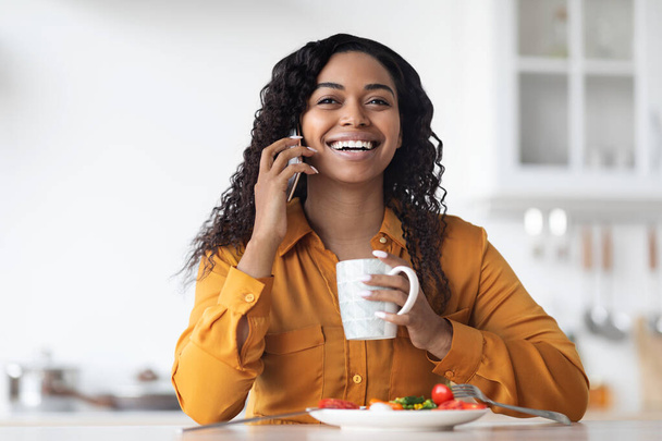 Cheerful black woman talking on phone while having breakfast - Фото, изображение