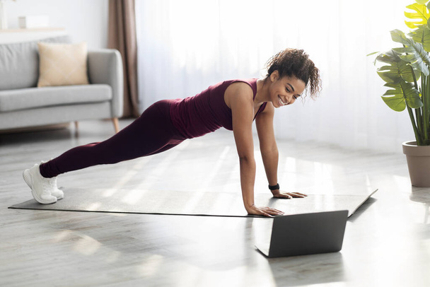 Positive slim african american woman exercising at home, using computer - Zdjęcie, obraz
