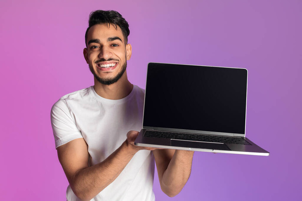 Hombre árabe milenario sonriente mostrando computadora portátil con pantalla negra vacía en luz de neón, maqueta para sitio web o anuncio - Foto, Imagen
