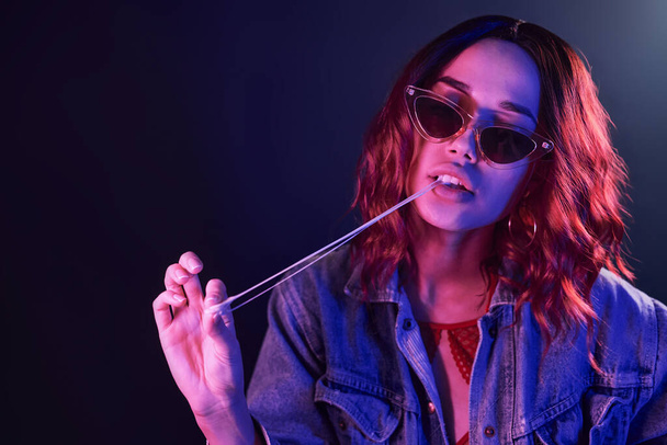 Portrait of young girl in glasses with bubble gum in red and blue neon in studio. - Foto, Bild