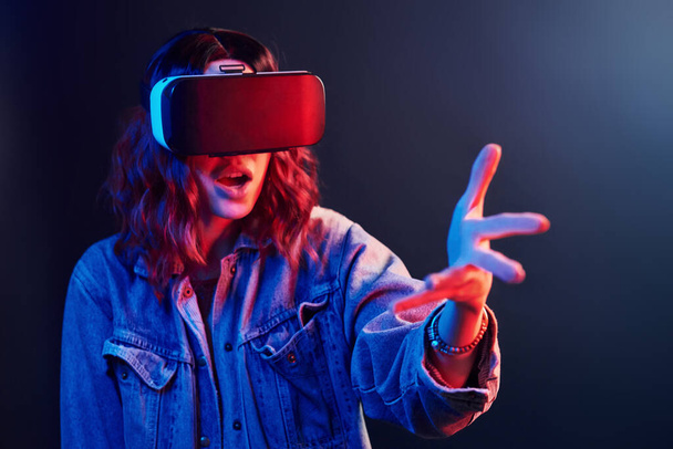 Facial expression of young girl with virtual reality glasses on head in red and blue neon in studio. - Foto, afbeelding
