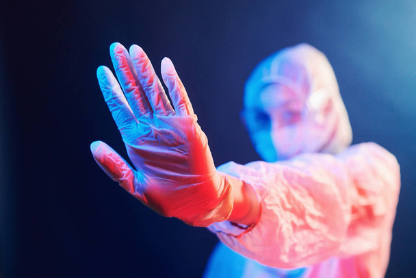 Nurse in mask and white uniform standing in neon lighted room and showing stop sign. - 写真・画像