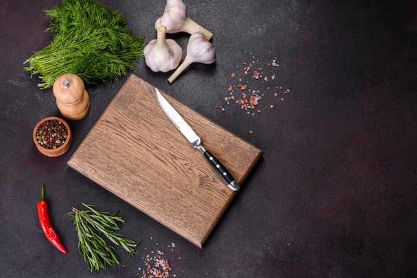 A wooden cutting board with spices, herbs, cherry tomatoes and salt on a black concrete background. Cooking at home - Zdjęcie, obraz