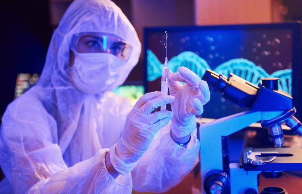 Медсестра в масці і білій формі, тримаючи шприц і сидячи в неоновій освітленій лабораторії з комп'ютером і медичним обладнанням в пошуках вакцини Коронавіруса. - Фото, зображення