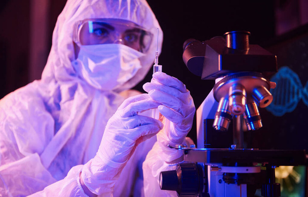Infermiera in maschera e uniforme bianca, in siringa e seduta in laboratorio illuminato al neon con computer e attrezzature mediche alla ricerca di vaccino Coronavirus. - Foto, immagini