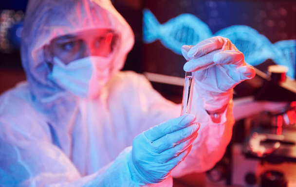 Enfermera en máscara y uniforme blanco, sosteniendo tubo con líquido y sentada en laboratorio con luz de neón con computadora y equipo médico en busca de vacuna contra el Coronavirus. - Foto, imagen