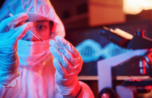 Hoitaja naamio ja valkoinen yhtenäinen, tilalla putki nesteellä ja istuu neon valaistu laboratoriossa tietokoneen ja lääketieteelliset laitteet etsivät Coronavirus rokote. - Valokuva, kuva