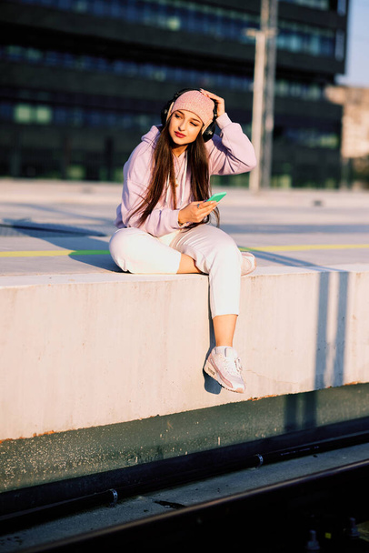 A teenage girl in urban wear sits on a railroad platform with headphones on and browses for a song on her phone. - Photo, image