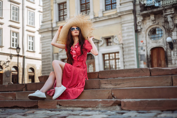 Καλοκαιρινή μόδα Κομψό κορίτσι με κόκκινο πουά φόρεμα, ψάθινο καπέλο και λευκά sneakers που κάθονται στις σκάλες. - Φωτογραφία, εικόνα