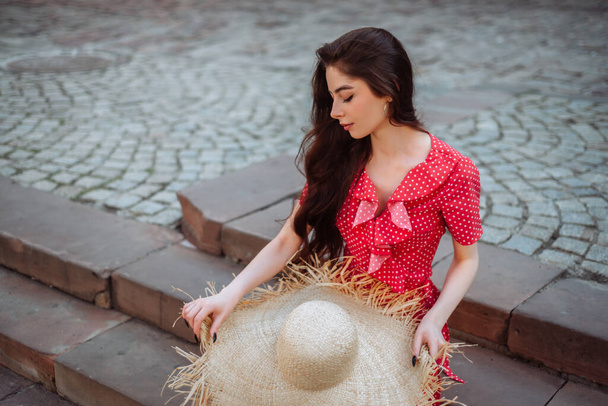 Ropa de verano: sombrero de paja y vestido rojo de lunares. Chica elegante sentada en las escaleras de la plaza de la ciudad. Retrato de verano - Foto, imagen