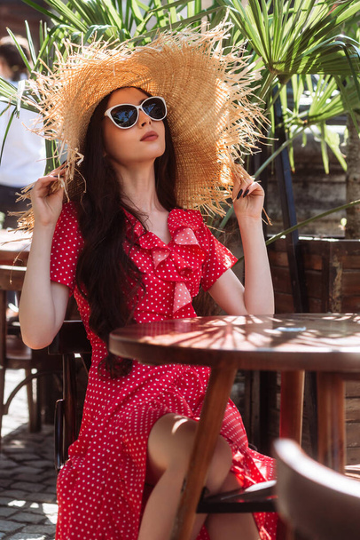 Girl in sunglasses, straw hat and red polka-dot dress relax on the summer terrace. Summertime portrait of young woman - Photo, Image