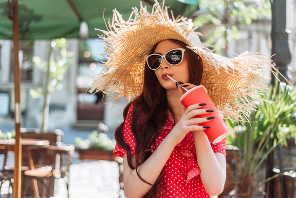 Menina elegante em óculos de sol, chapéu de palha e vestido de bolinhas vermelhas relaxar no terraço com bebida fria. Retrato de verão de mulher jovem - Foto, Imagem