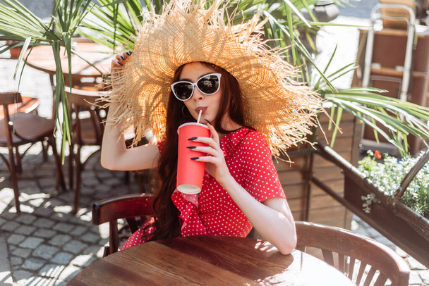 Retrato de verão da jovem relaxada em óculos de sol e chapéu de palha com bebida gelada sentada no terraço ensolarado - Foto, Imagem