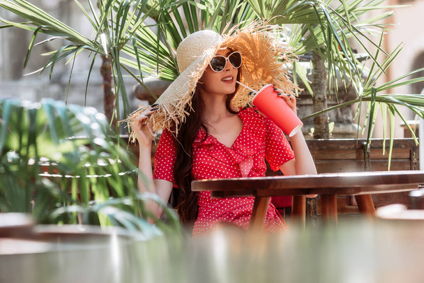 Стильна дівчина в сонцезахисних окулярах, солом'яний капелюх і червона сукня-полька розслабляються на терасі з холодним напоєм. Літній портрет молодої жінки
 - Фото, зображення