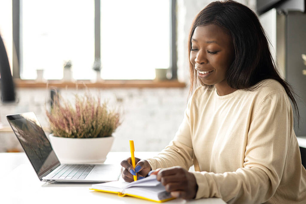 A beautiful African-American woman in casual wear is using laptop computer sitting in home office - Photo, Image