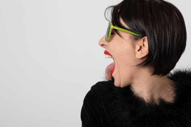 Portrait of elegant woman dressed in black on a white background. Aggressive and provocative profile. She has green sunglasses. Her mouth is open and she sticks her tongue out as if she is disgusted or screaming. - Foto, Bild