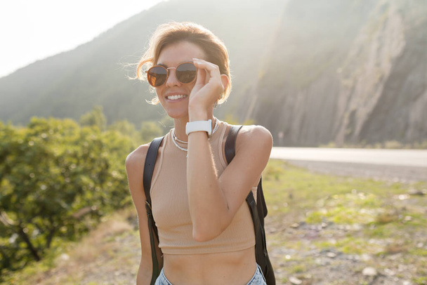 Outdoor portrait of charming stylish european girl with light hair wearing sunglasses and beige t-shirt with backpack is traveling among mountains in sunlight in sunny day. High quality photo - Fotó, kép