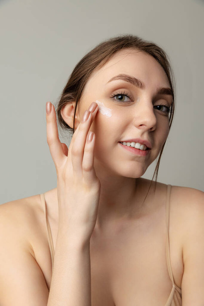 Portrait of beautiful young girl with perfect smooth skin applying face moisturizing cream isolated over grey studio background - Valokuva, kuva