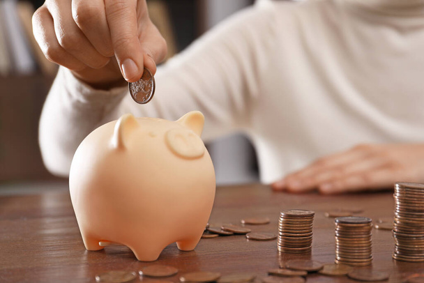 Woman putting coin into piggy bank at wooden table indoors, closeup - Photo, image