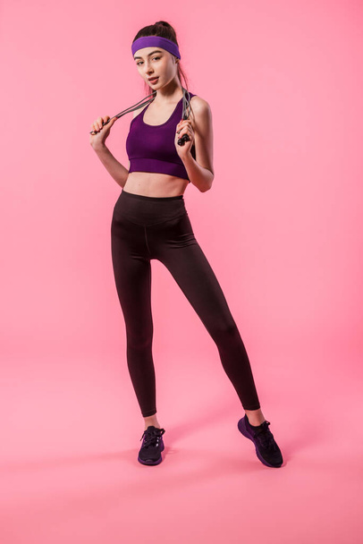 Sportlerin in Leggings und Top mit Springseil auf rosa Hintergrund. Sport, aktiver, gesunder Lebensstil. Athletische Fitness-Mädchen in voller Länge Porträt - Foto, Bild