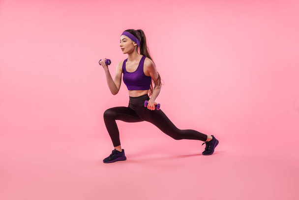 Athletic girl training with dumbbells pumping up muscles of hands and legs on pink background. Sport, active healthy lifestyle - 写真・画像
