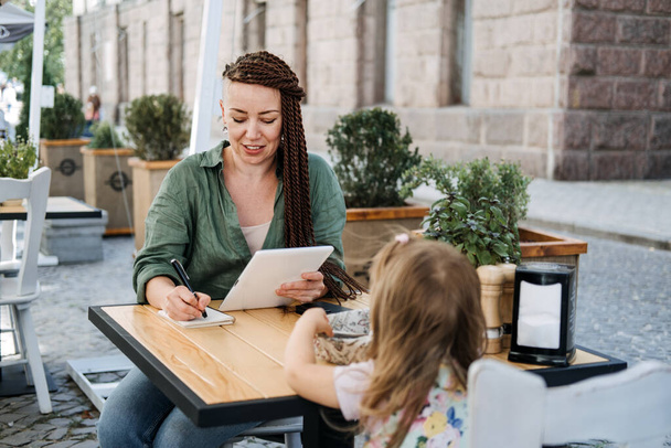 Freelance Parents. Busy working mom with cell phone and tablet working in street cafe with baby toddler daughter. Multitasking Freelancer mom and her baby daughter working together outdoors. - Foto, Imagem