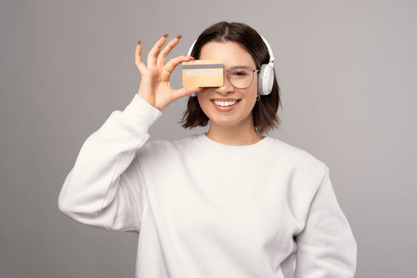 Smiling woman is holding a credit card and hiding behind while wearing headphones. - Foto, afbeelding