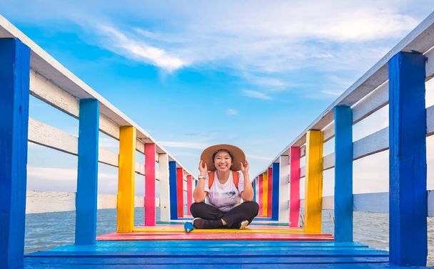 Perspective view of happy Asian woman smiling while sitting on colorful wooden bridge at sea viewpoint against cloud on blue sky background  - Foto, imagen