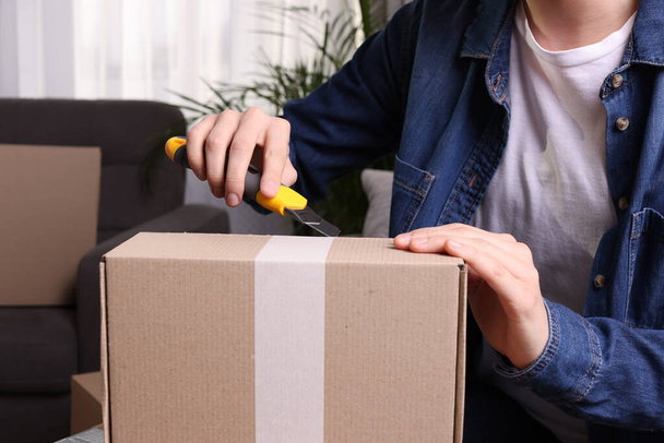 Young woman using utility knife to open parcel at home, closeup - Foto, imagen