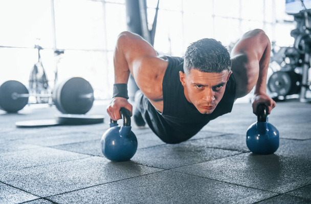 Strong young man in sportive clothes doing push ups in gym by using dumbbells. - Foto, Imagem
