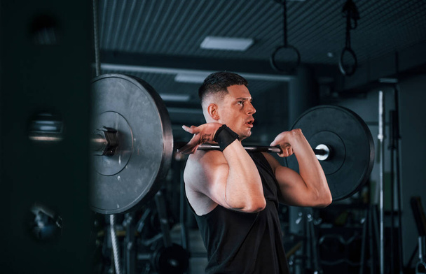 Strong young man in sportive clothes doing exercises with heavy weights in the gym. - 写真・画像