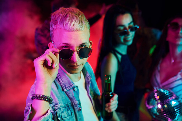 Hipster guy in sunglasses and with bottle of alcohol posing for camera in front of young people that having fun in night club with colorful laser lights. - Fotoğraf, Görsel