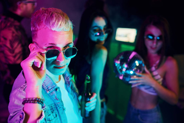 Hipster guy in sunglasses and with bottle of alcohol posing for camera in front of young people that having fun in night club with colorful laser lights. - Foto, afbeelding