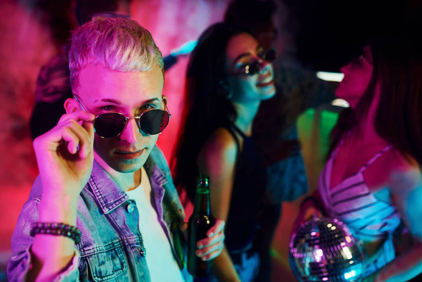 Hipster guy in sunglasses and with bottle of alcohol posing for camera in front of young people that having fun in night club with colorful laser lights. - Foto, Bild