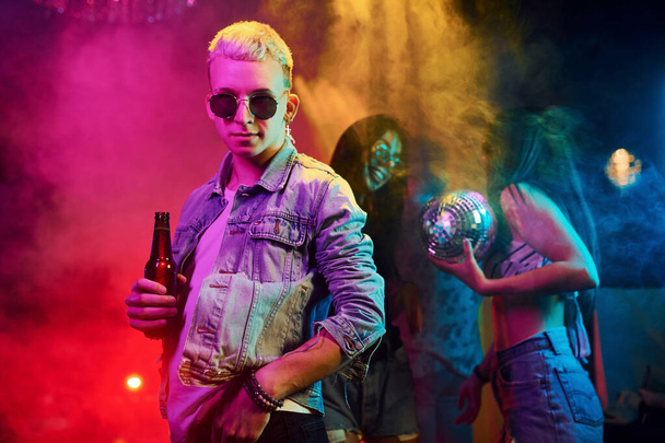 Hipsterski facet w okularach przeciwsłonecznych i z butelką alkoholu pozujący do kamery przed młodymi ludźmi, którzy bawią się w nocnym klubie z kolorowymi światłami laserowymi. - Zdjęcie, obraz