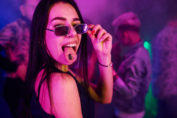 Young girl holding drug pills on togue in front of young people that having fun in night club with colorful laser lights. - Foto, immagini