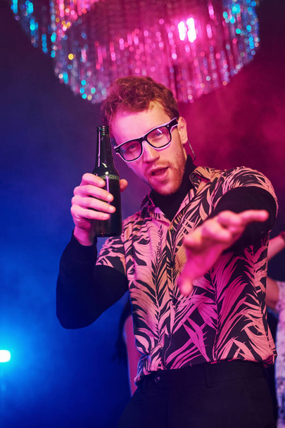 Guy in glasses drinks beer. Young people is having fun in night club with colorful laser lights. - Fotó, kép