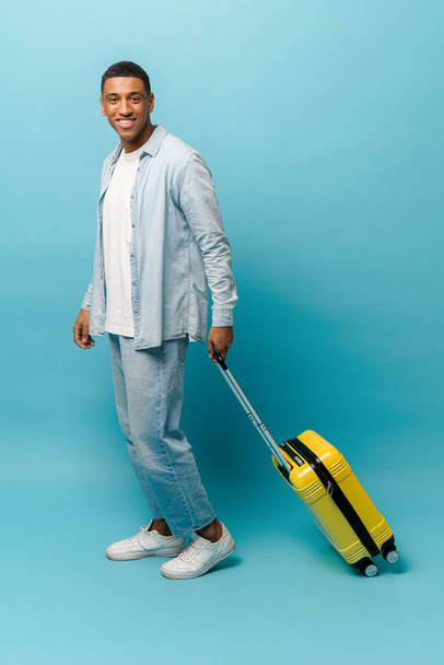 Счастливый афроамериканец в повседневной рубашке и джинсах ходит с желтым чемоданом - Фото, изображение