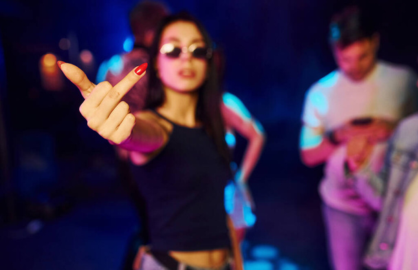 Woman in glasses shows middle finger. Young people is having fun in night club with colorful laser lights. - Photo, image