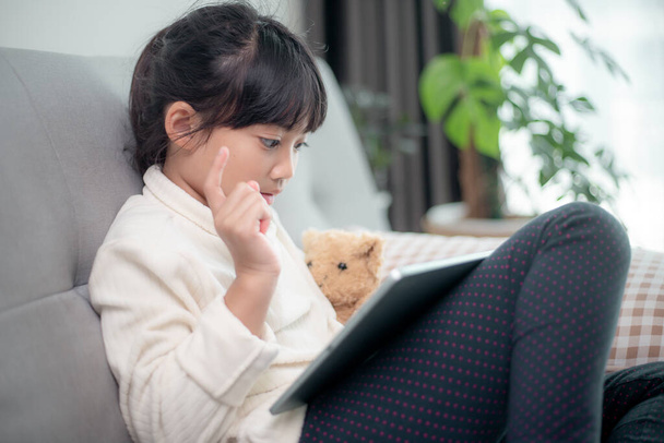 Κοριτσάκι που χρησιμοποιεί tablet παιχνίδι στο διαδίκτυο, Παιδί που κάθεται στον καναπέ βλέποντας ή μιλώντας με ένα φίλο σε απευθείας σύνδεση, Παιδί χαλαρώνοντας στο σαλόνι το πρωί, Παιδιά με την έννοια της νέας τεχνολογίας - Φωτογραφία, εικόνα