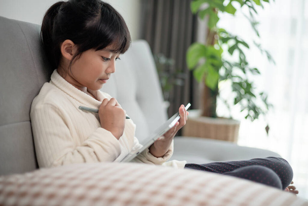 İnternette tablet kullanan küçük bir kız kanepede oturan ya da arkadaşıyla internetten konuşan bir çocuk, sabah oturma odasında dinlenen çocuk, yeni teknoloji konsepti olan çocuklar. - Fotoğraf, Görsel