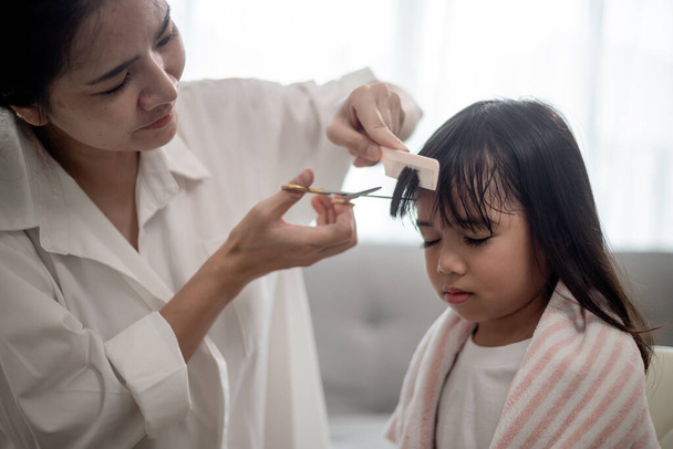 Mère asiatique coupe les cheveux de sa fille dans le salon à la maison tout en restant à la maison à l'abri de Covid-19 Coronavirus pendant le confinement. Concept d'auto-quarantaine et de distance sociale. - Photo, image