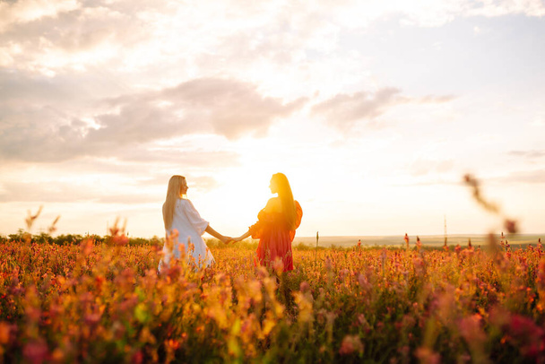 Portrait of two young woman in stylish dress posing in the blooming field. Nature, vacation, relax and lifestyle. Fashion concept. - 写真・画像