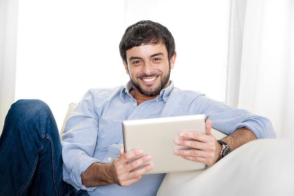 Young attractive Hispanic man at home on white couch using digital tablet or pad - Fotoğraf, Görsel