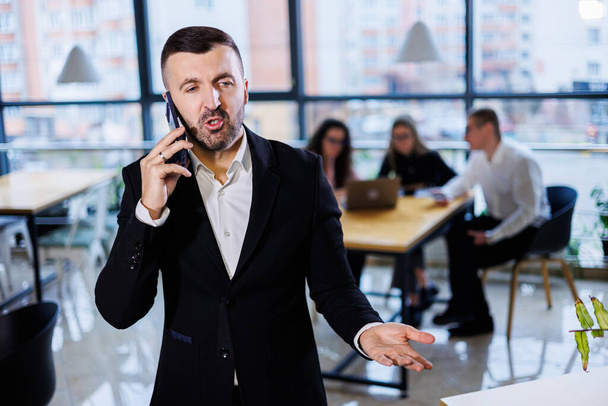 Красивый молодой бизнесмен в современном костюме с помощью мобильного телефона, успешный мужчина разговаривает по телефону в офисе - Фото, изображение